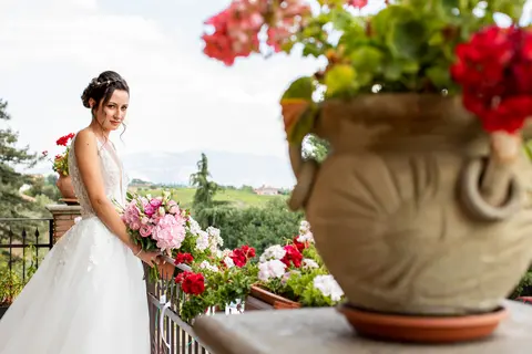 La sposa Erika in abito bianco tiene un bouquet di fiori su un balcone con vasi di fiori in primo piano.