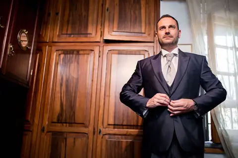 Lo sposo Daniele si abbottona il completo in una stanza con armadi in legno.