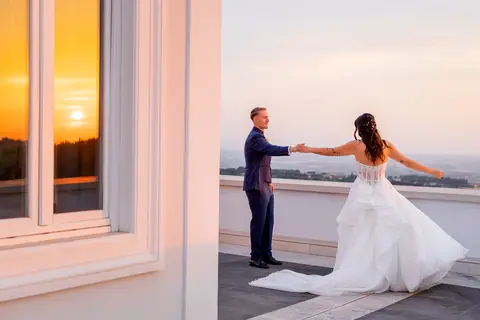 Il sole che tramonta riflesso sulla finestra della villa e lo sposo che porta la sposa per mano sulla terrazza