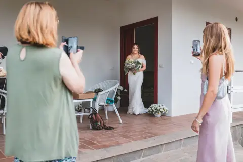 Un'invitata riprende con il telefono l'uscita della sposa dal portico